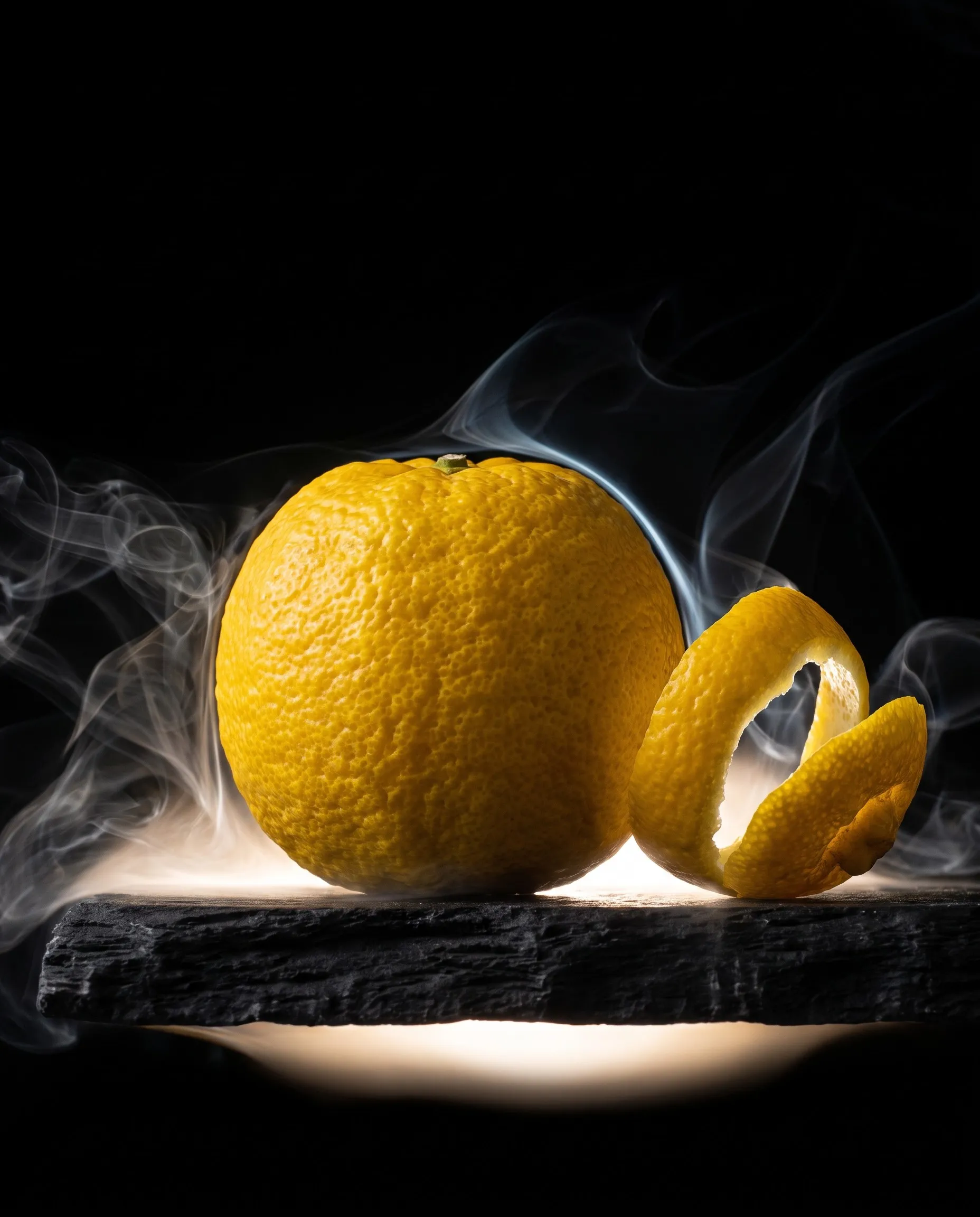 Buah yuzu kuning dengan kulit yang terkupas melingkar di atas batu hitam, dikelilingi asap tipis